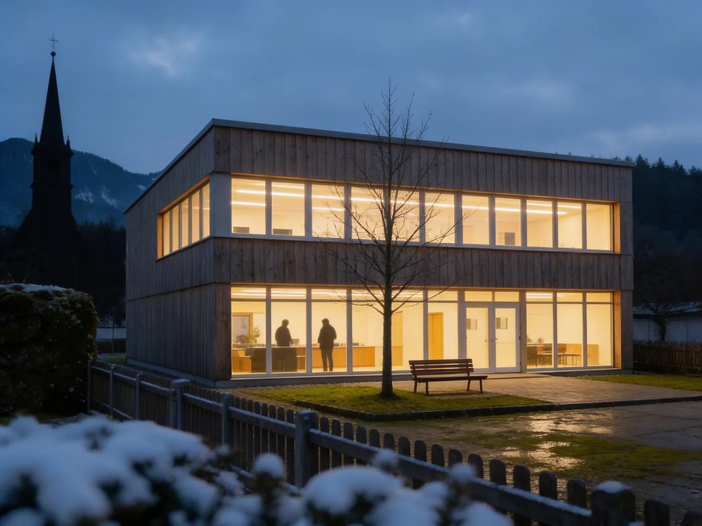 Municipal office lit up at dusk in winter, two people inside, church tower behind